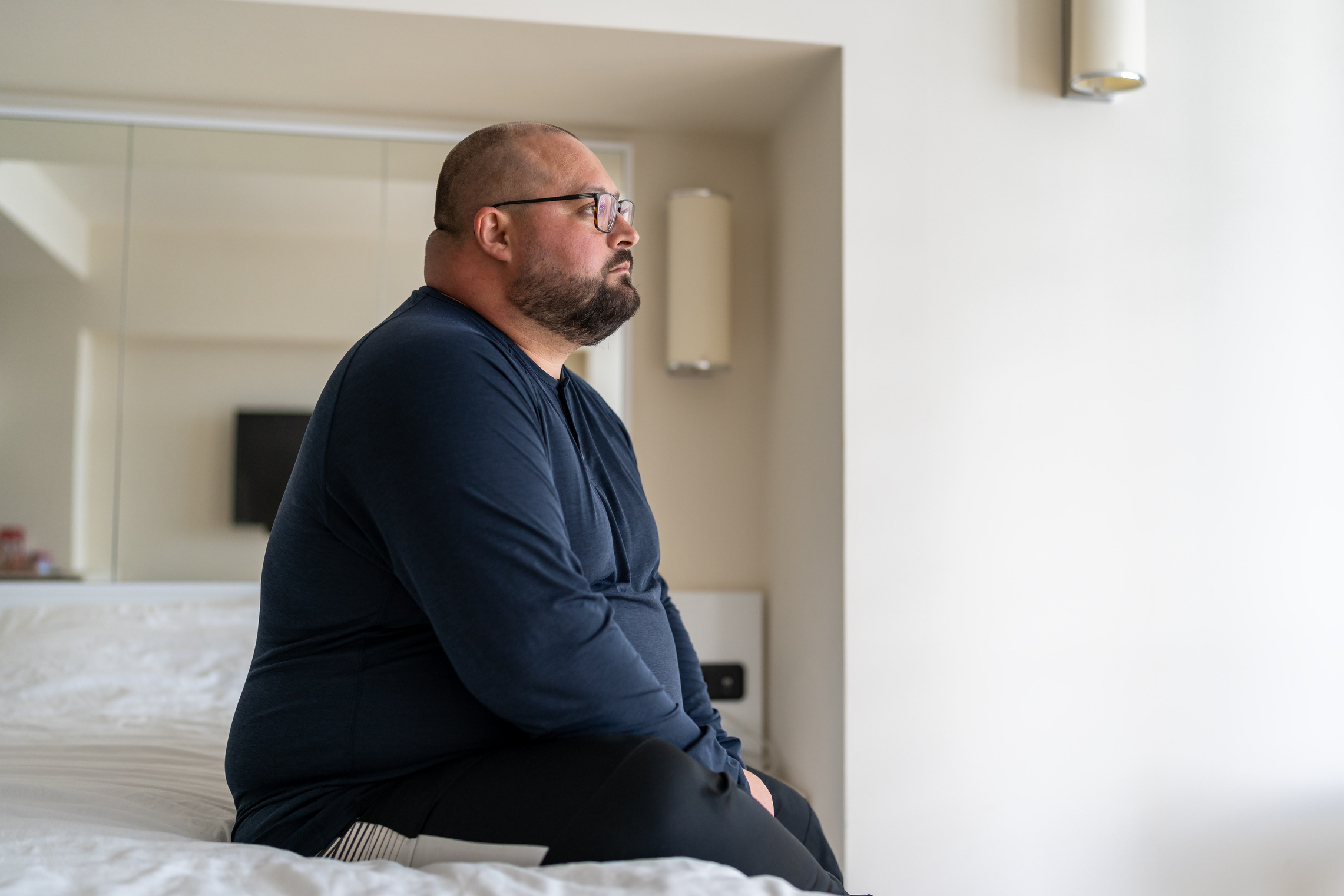 Sad fat lonely man sitting on bed in hotel and looking out window, thinking about life problems